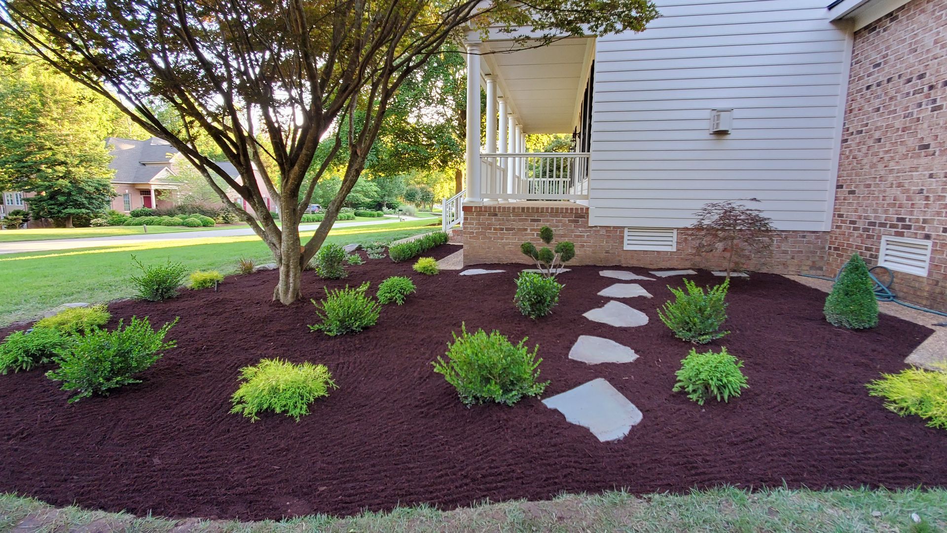 Front yard landscaping with small shrubs, stepping stones, and a tree beside a house porch