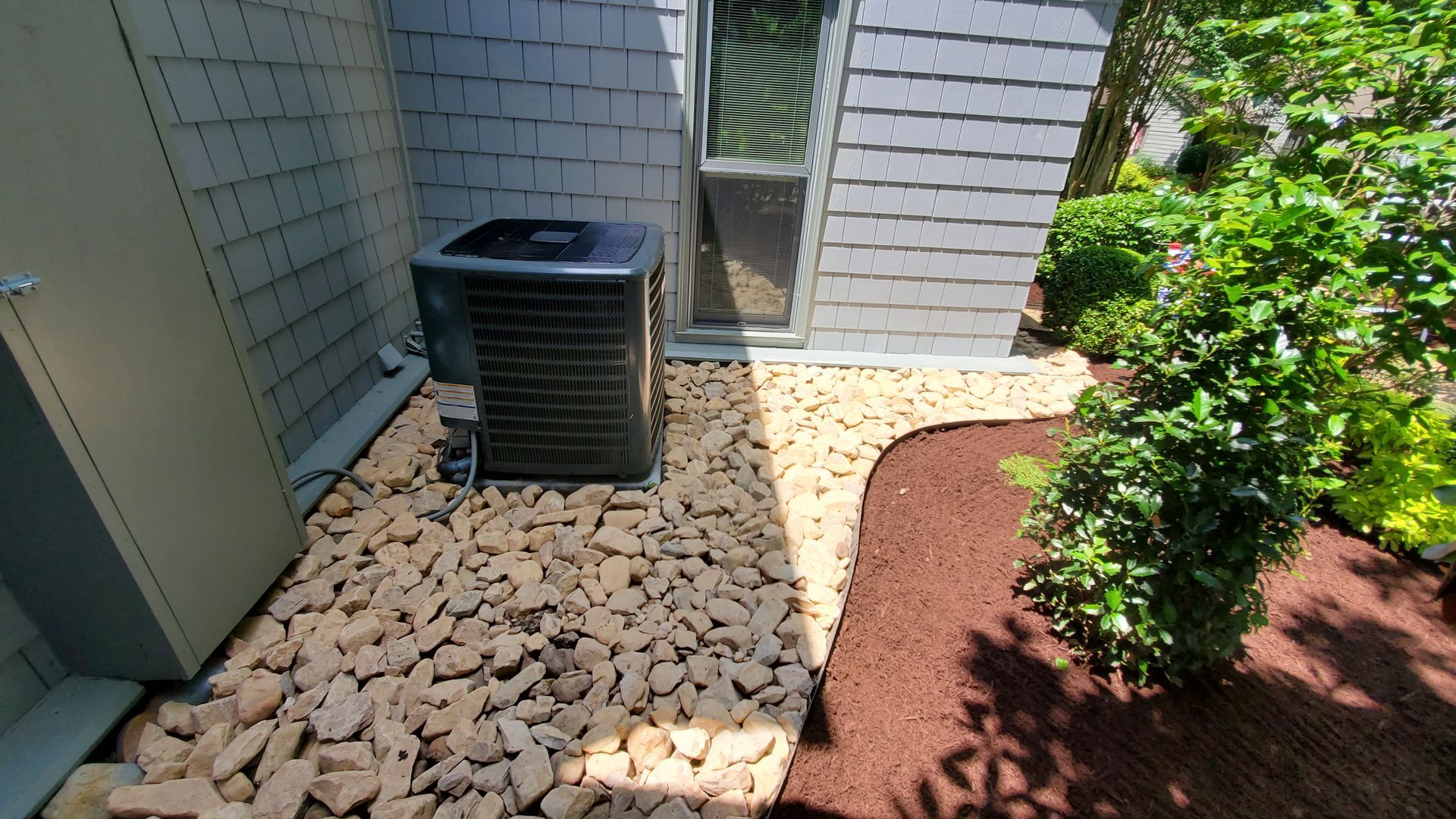 Outdoor HVAC unit beside a white house wall, with white rocks, mulch, and green shrubs nearby