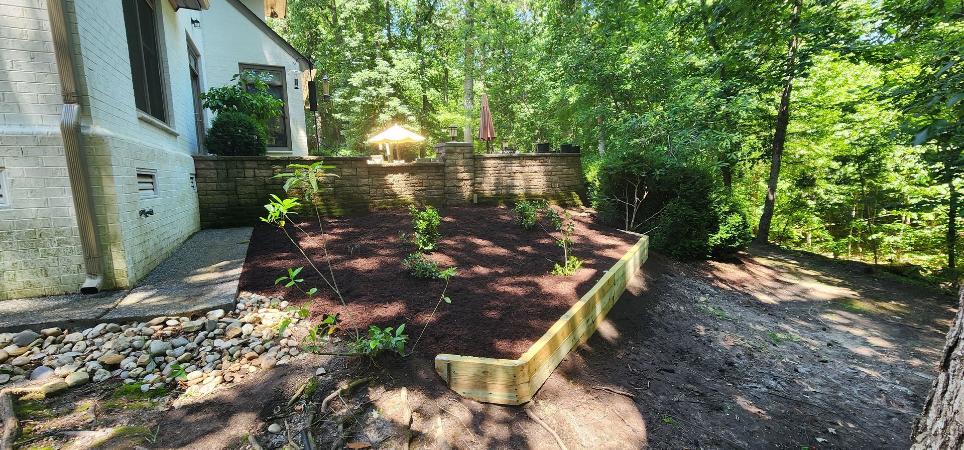 Mulched garden bed with low wood edging beside a house, surrounded by trees and gravel path