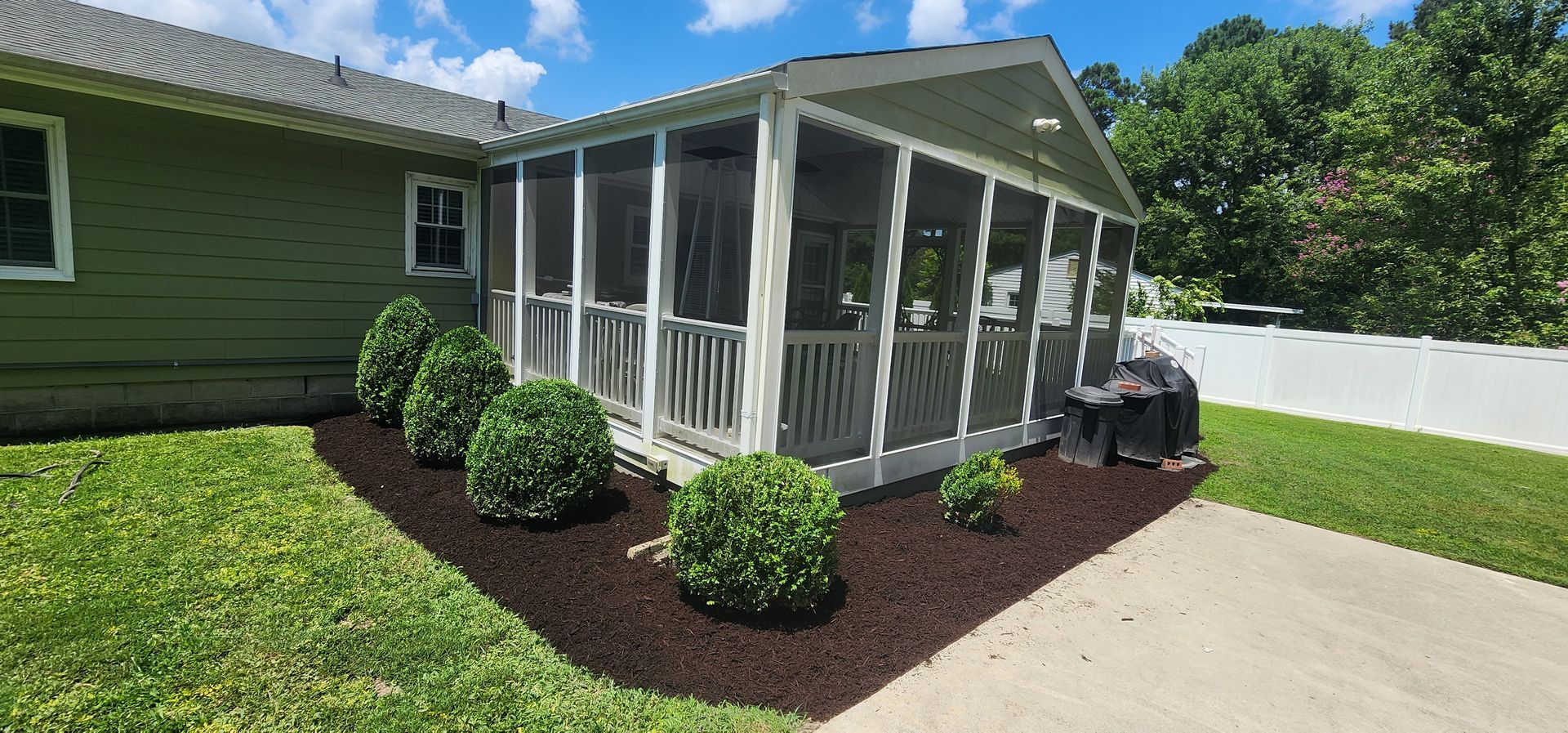 Screened patio beside green house, with mulched shrubs and a concrete driveway under bright sun