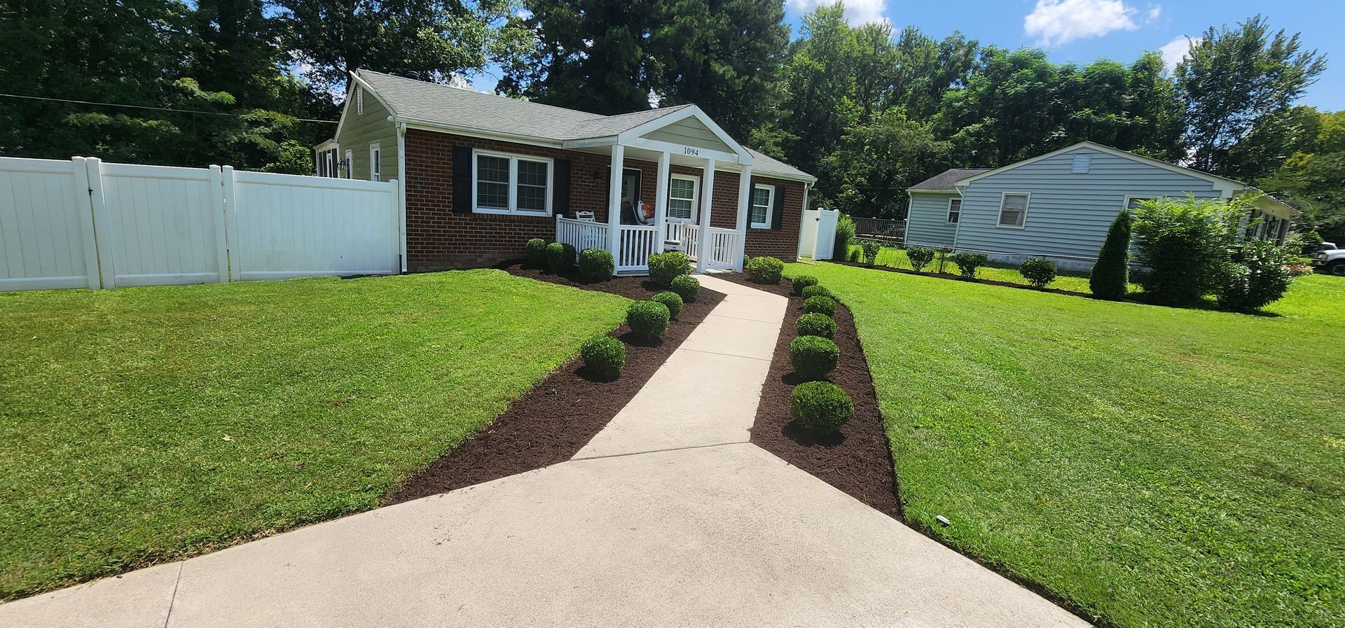Front yard walkway leading to a brick house with white trim, green lawn, and white fence under a blue sky