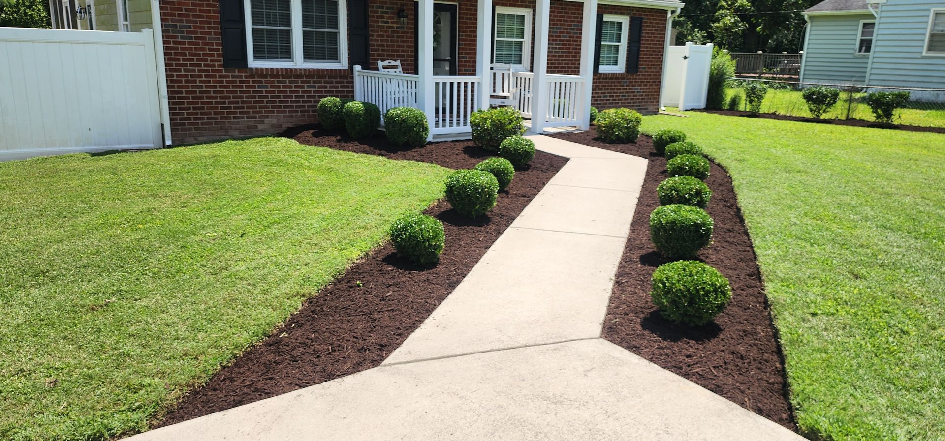 Landscaped garden with mulch beds, trees, and a concrete walkway beside a patio.