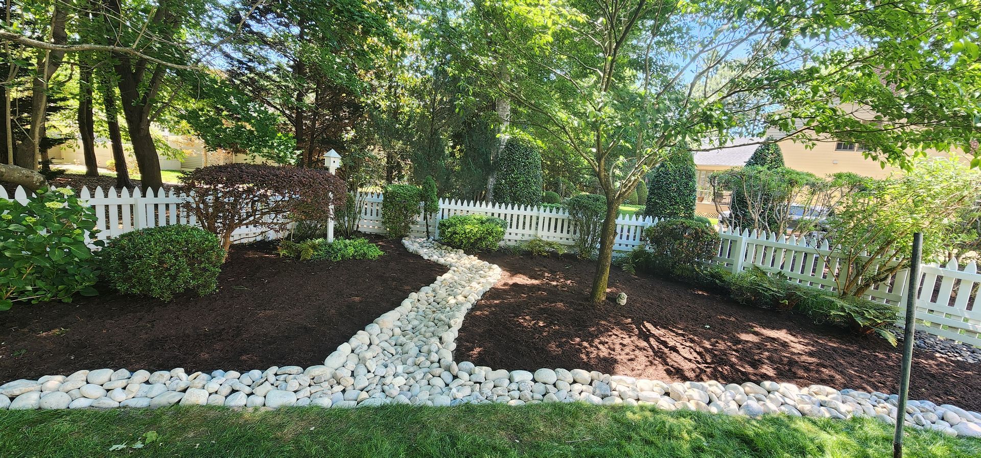 Garden path lined with white stones, mulch beds, and white picket fencing under trees in a sunny yard