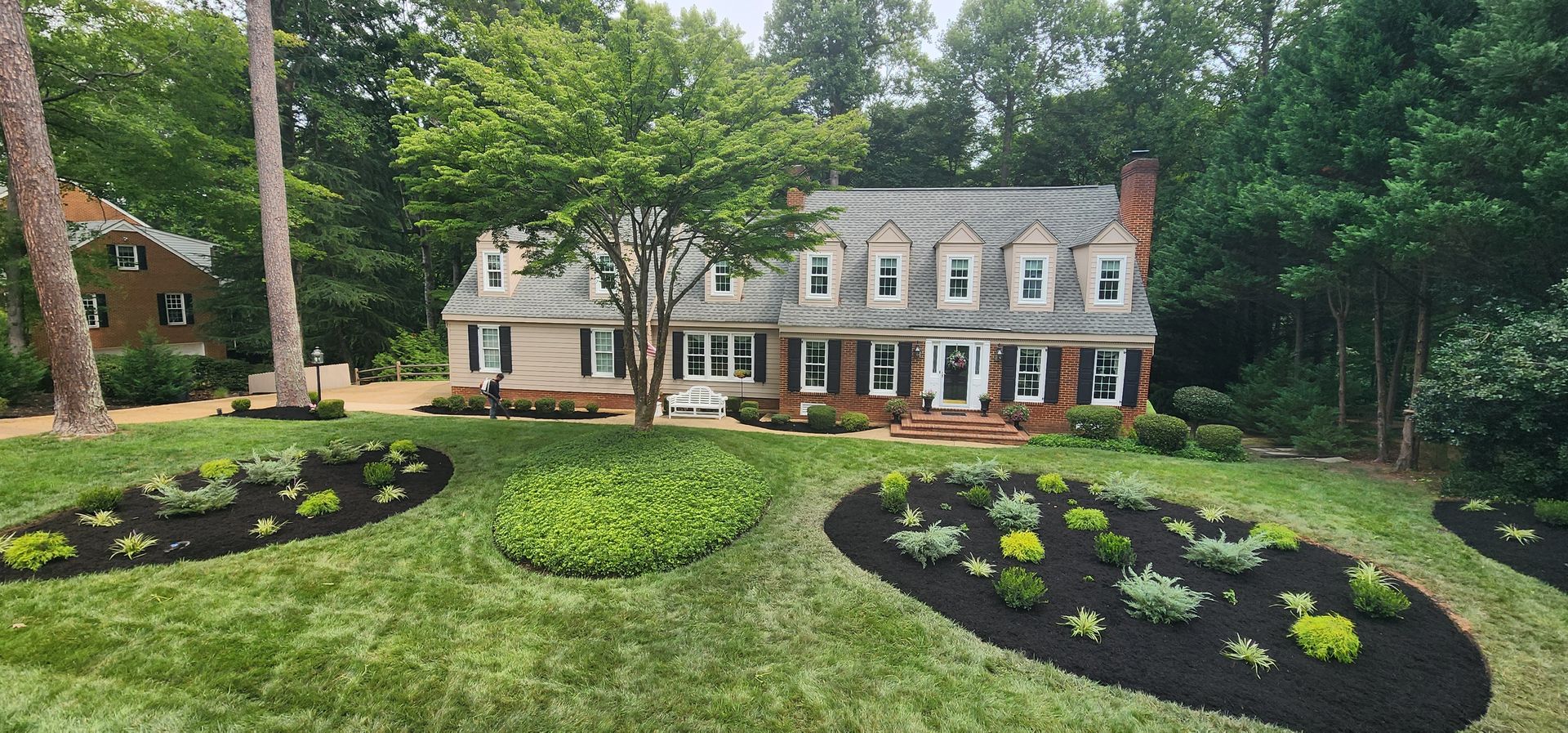 Landscape of a large beige house with manicured lawn and flower beds surrounded by trees