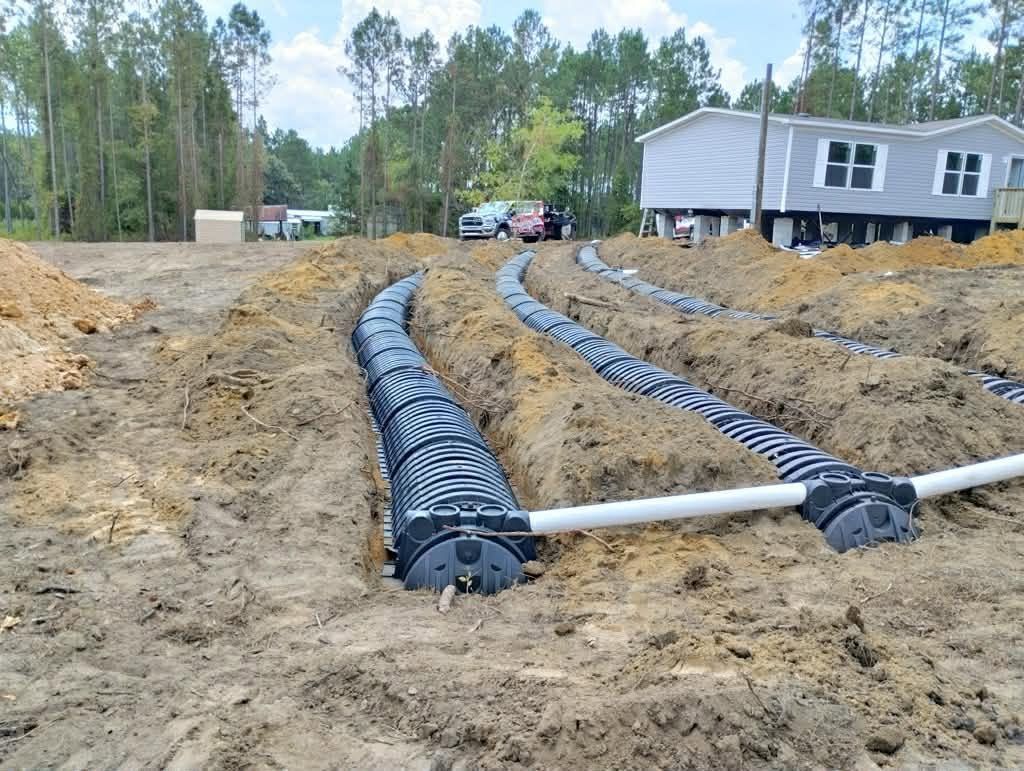 Septic drain field trenches with black perforated pipes beside a gray house in a wooded yard