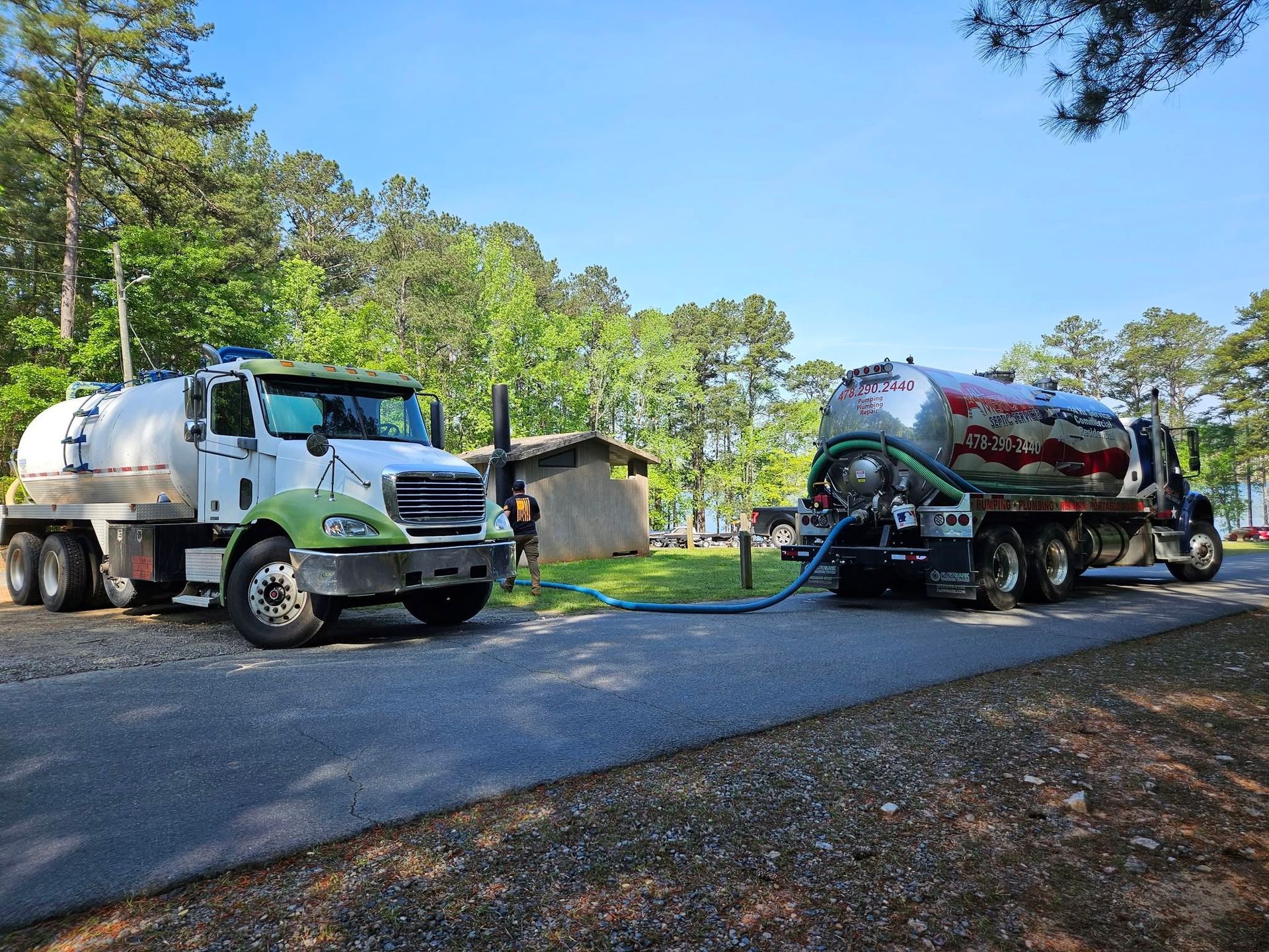 Two tanker trucks on a roadside in a wooded area under a clear blue sky