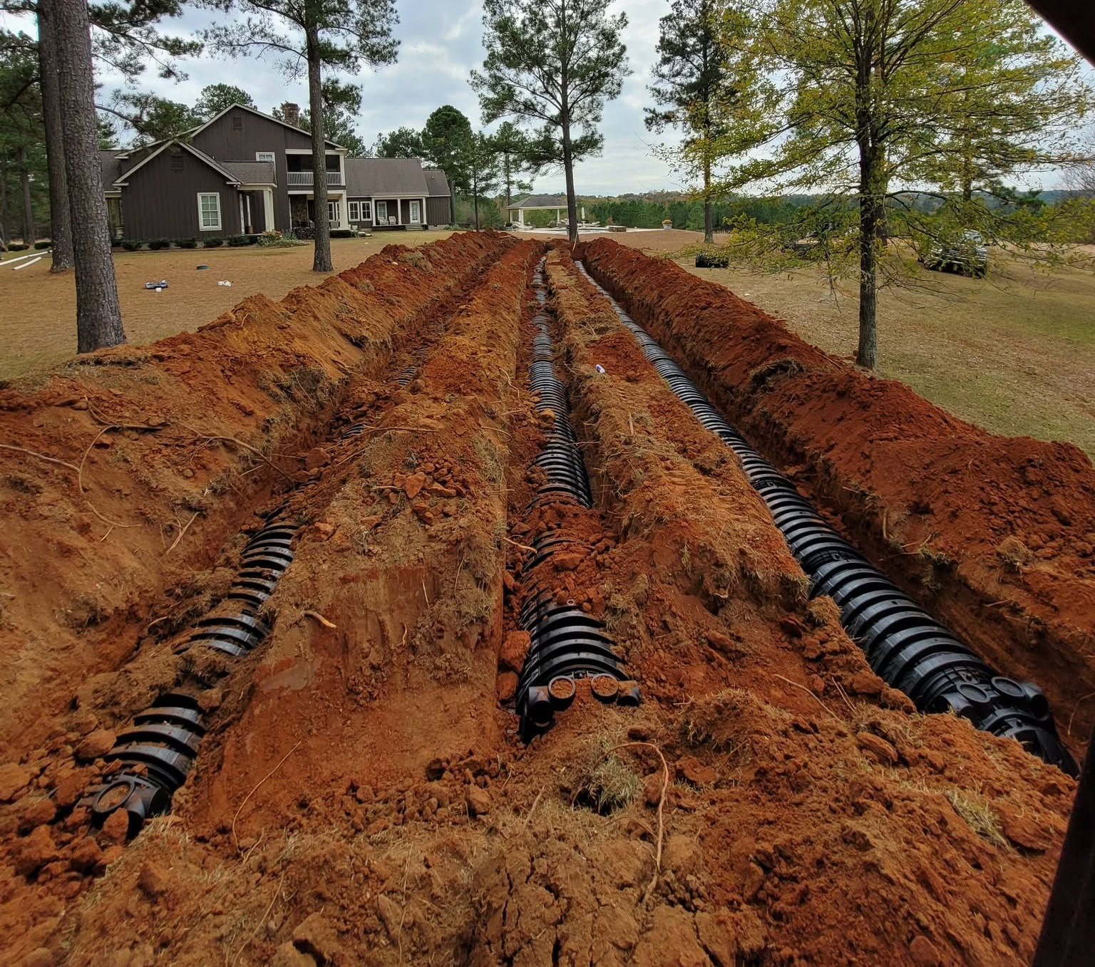 Construction trenches with black drainage pipes laid in red soil near a house and trees