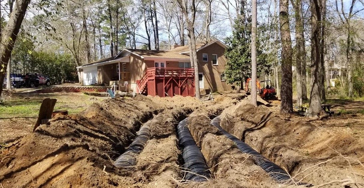Trenched yard with exposed black pipes leading to a house under construction among trees