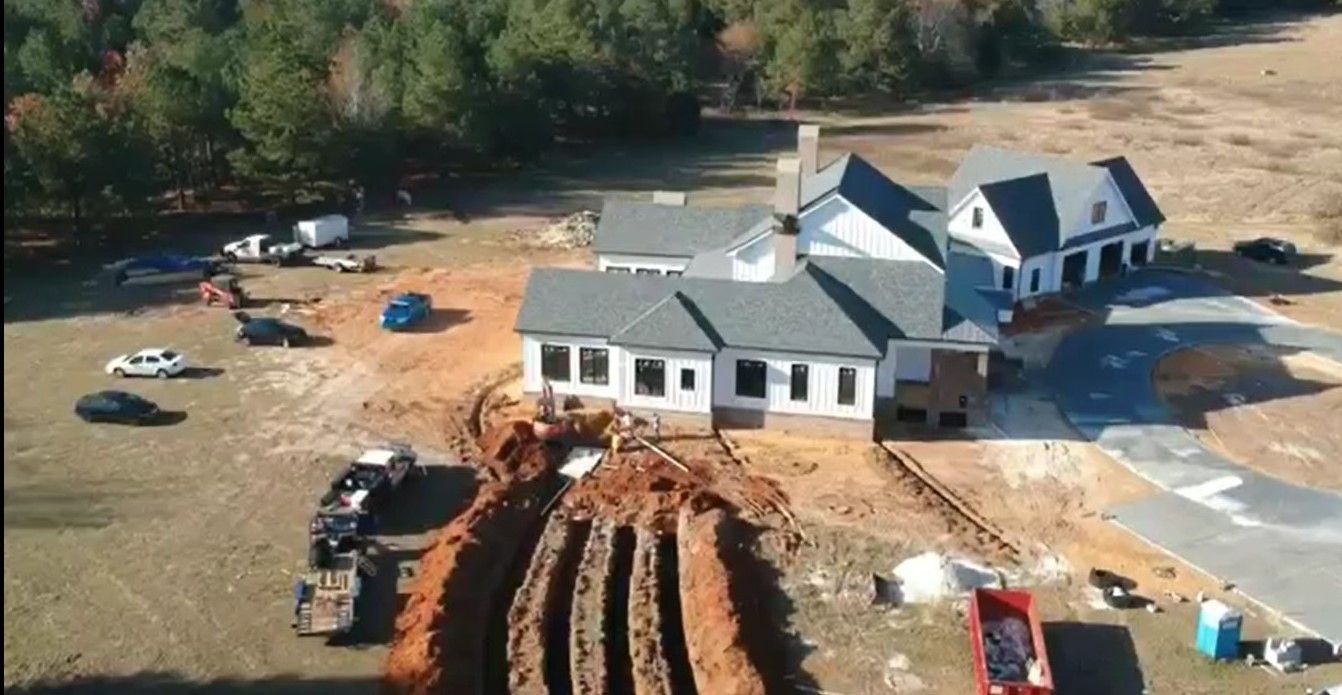 Aerial view of a large house under construction with dirt piles, trucks, and trees around it.