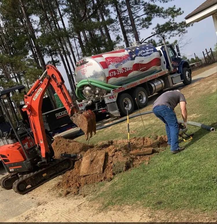 Excavator and worker digging a trench beside a tanker truck on a grassy roadside.