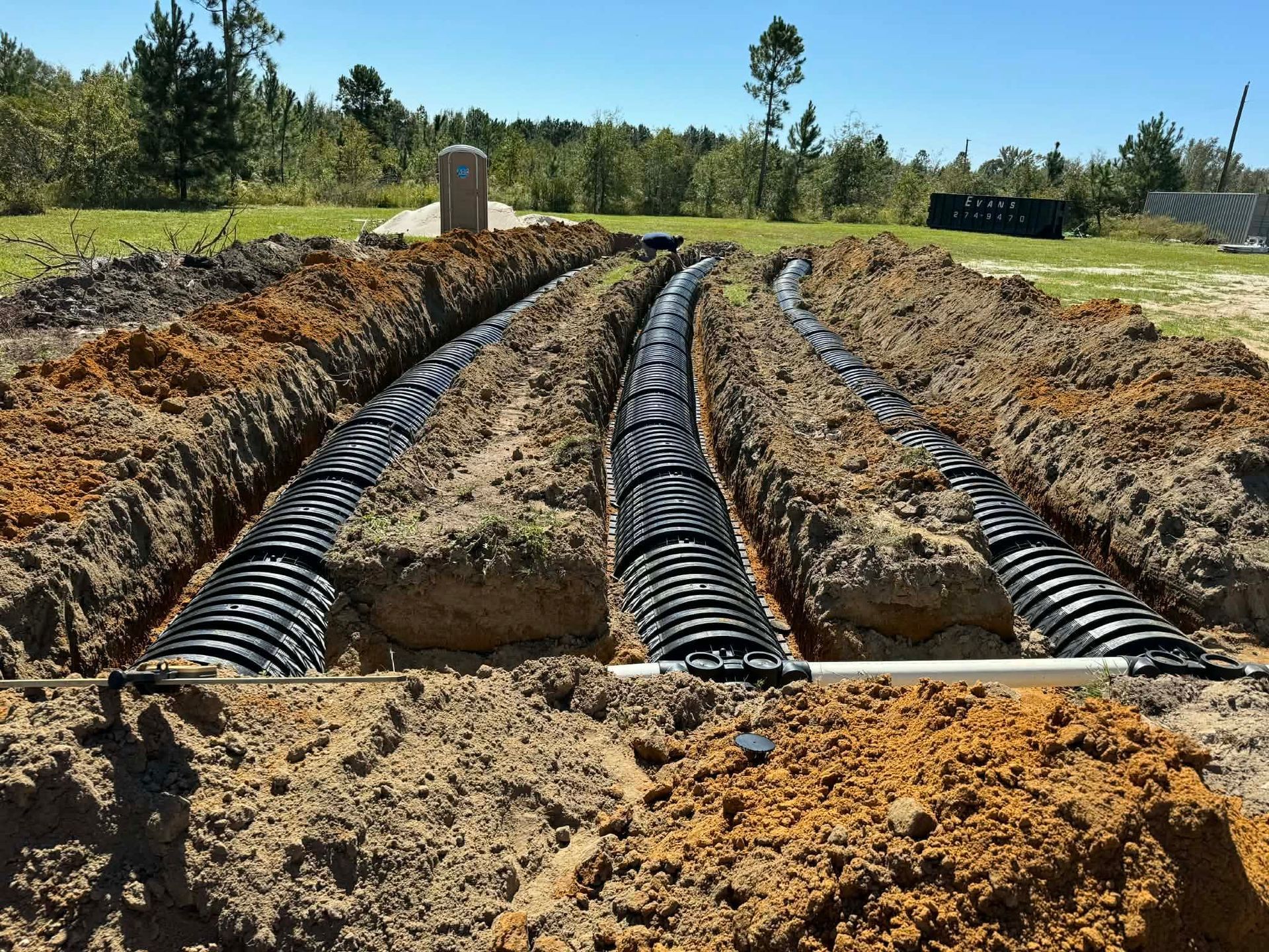 Trenched ground with black corrugated drainage pipes laid in parallel under a sunny grassy field.