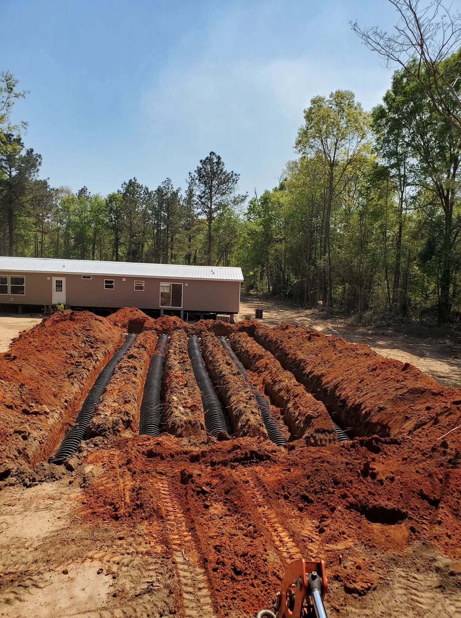 Red dirt foundation trenches beside a brick house under construction, with trees in the background