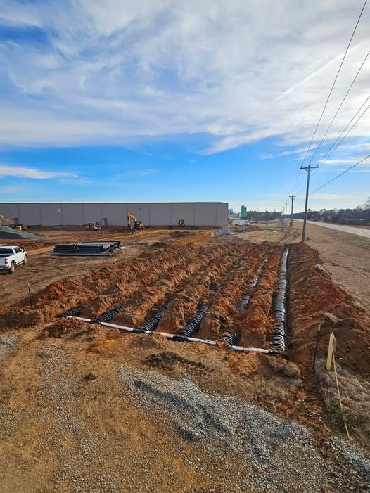 Roadside construction site with exposed dirt, pipes, a large warehouse, and power lines under a blue sky