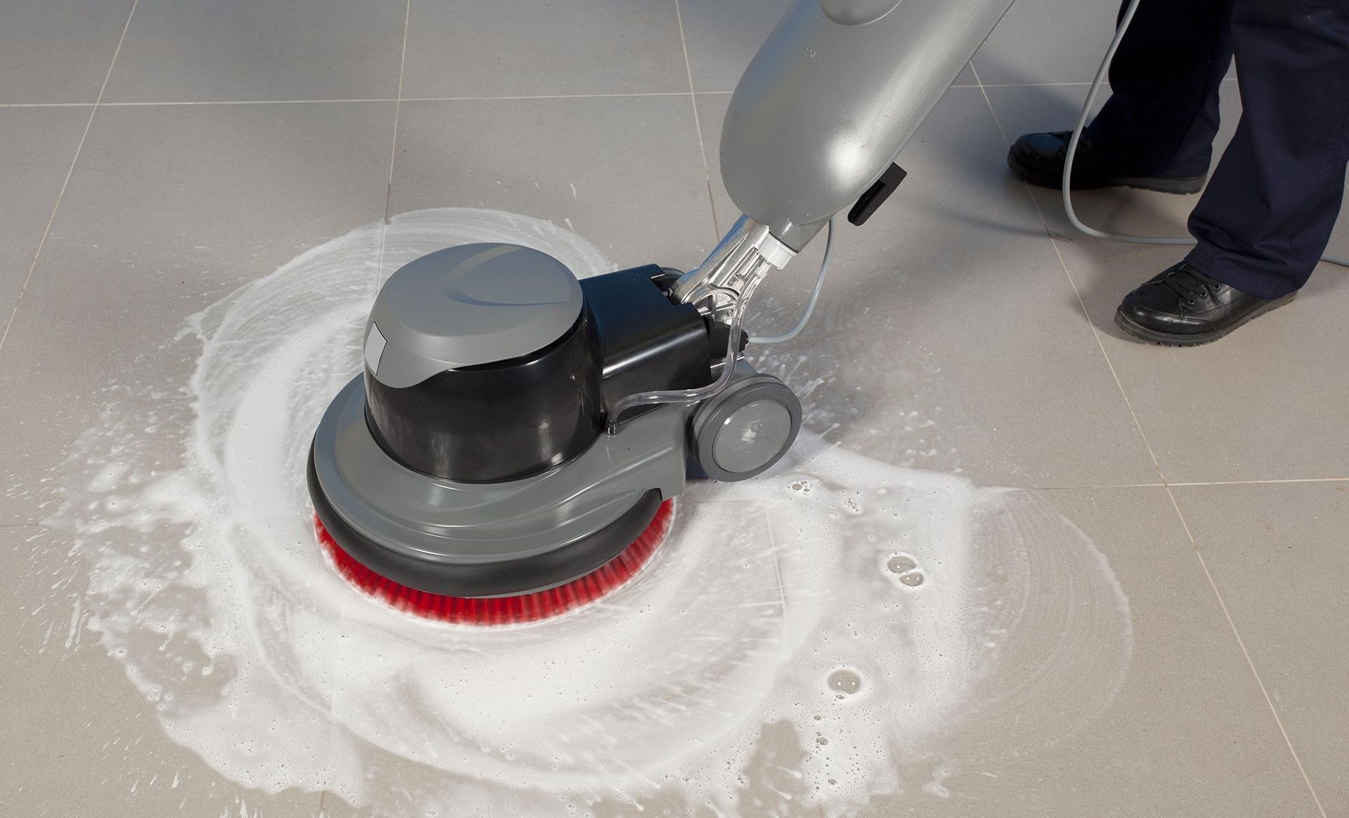 A person using a gray floor scrubber with a red brush, cleaning a tiled floor.
