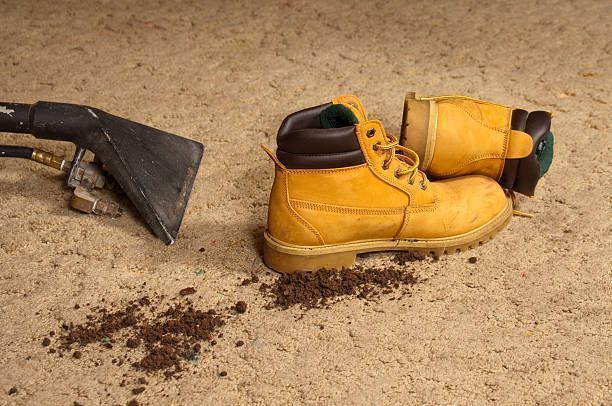 Yellow work boot and gloves next to a soil stain on tan carpet, with a carpet cleaning tool on the left.