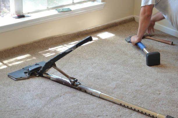 Man installing carpet with a knee kicker and stretcher near a window in a beige room.