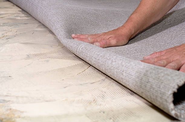 Person rolling out a gray carpet onto a light-colored surface with a visible grid pattern.