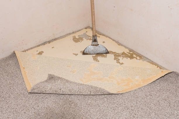 A person using a floor scraper to remove old carpet from a room corner.