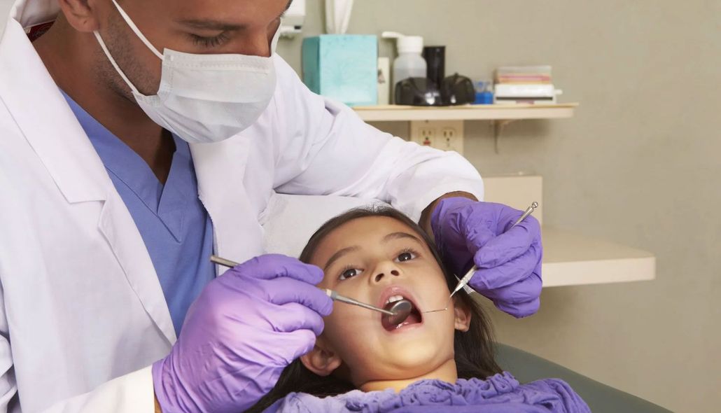 Dentist in mask and gloves examining a child’s open mouth in a clinic chair.