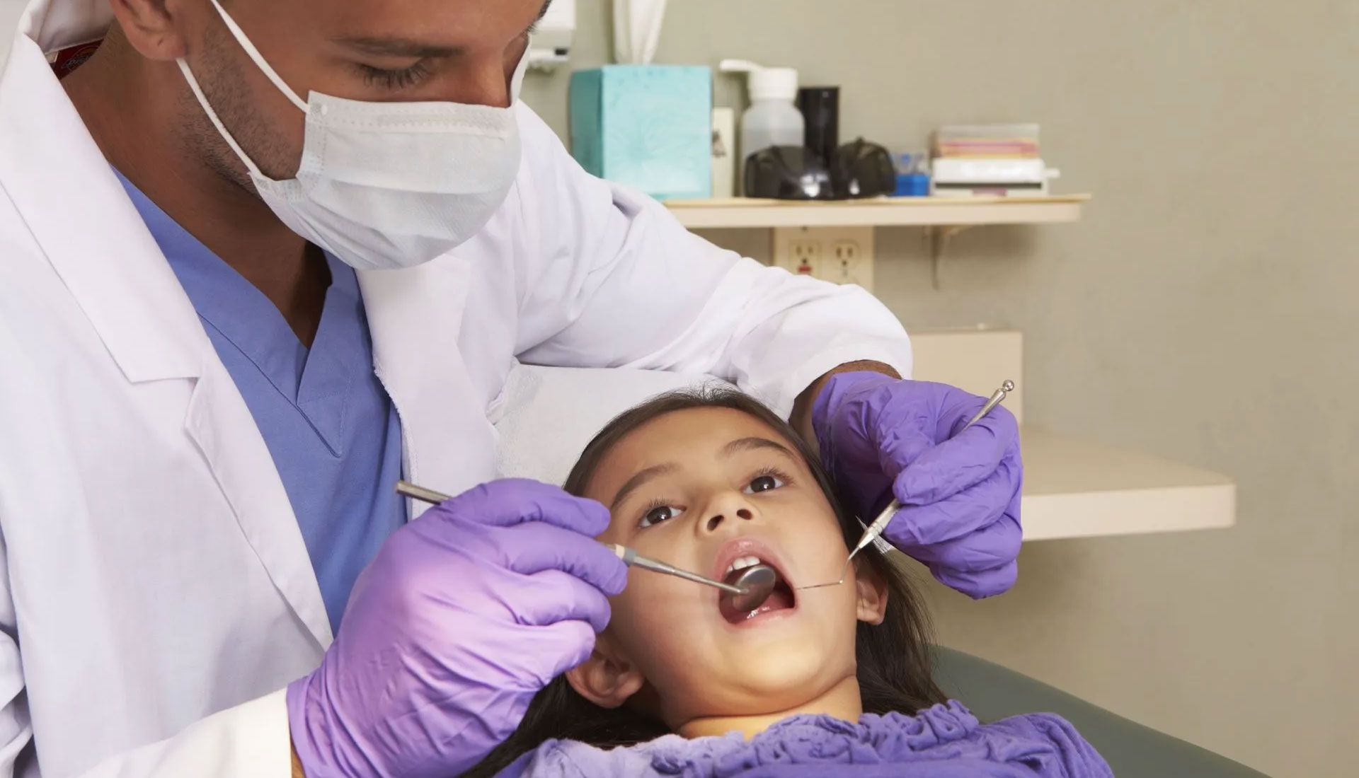 Dentist in mask and gloves examining a child’s open mouth in a clinic chair.