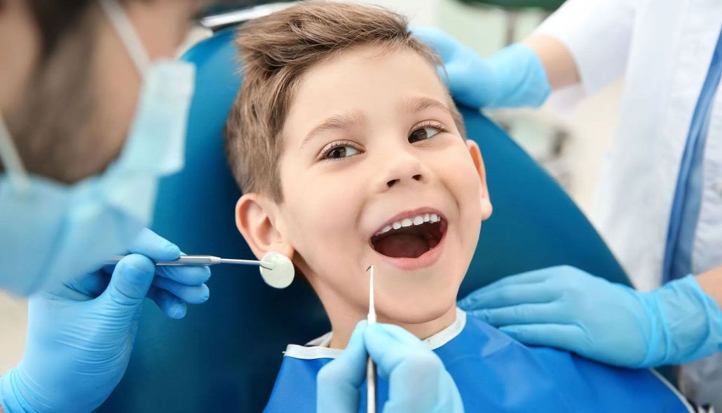 Child smiling during a dental checkup, with dentists wearing blue gloves and dental tools nearby.