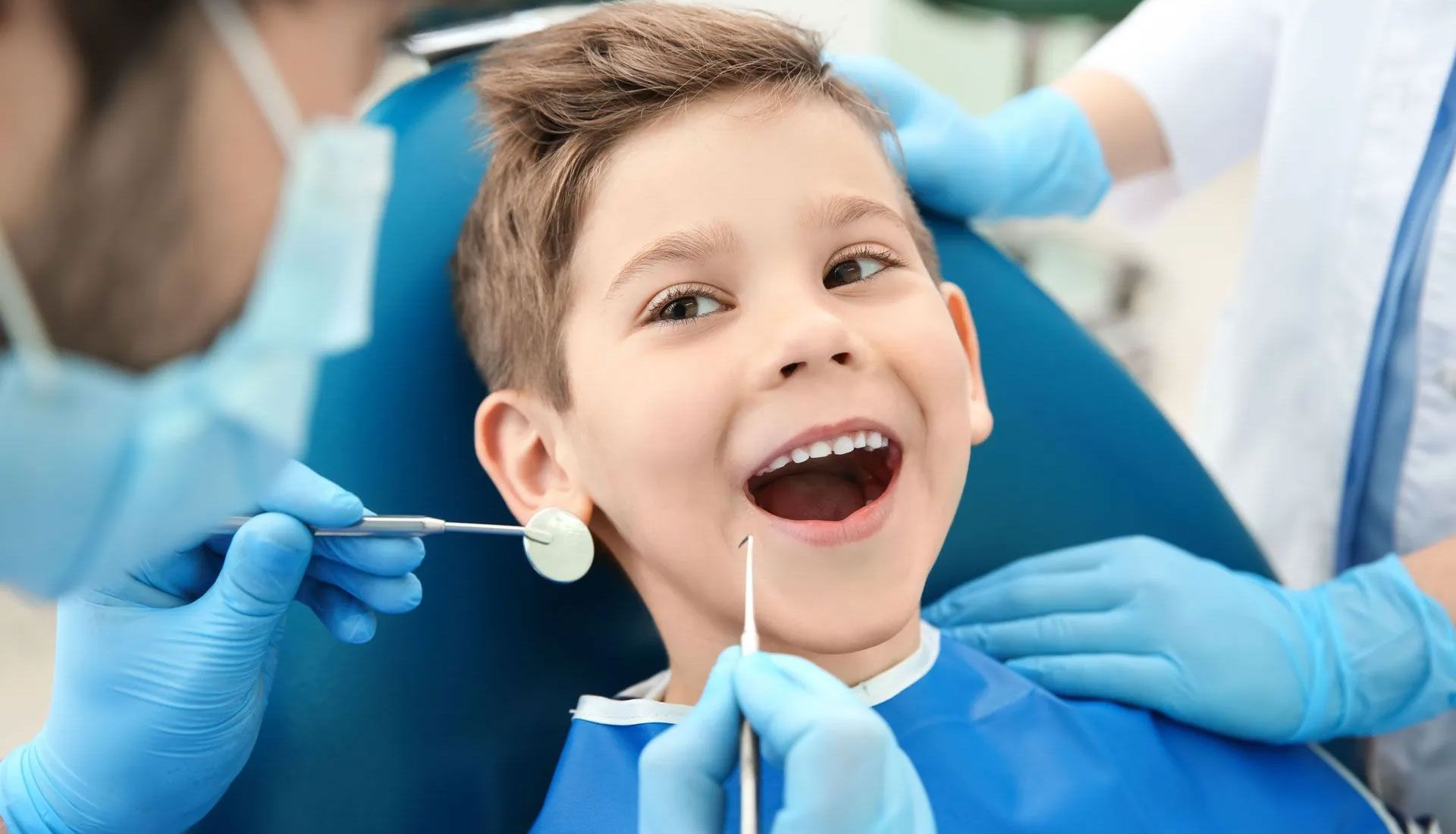 Child smiling during a dental checkup, with dentists wearing blue gloves and dental tools nearby.