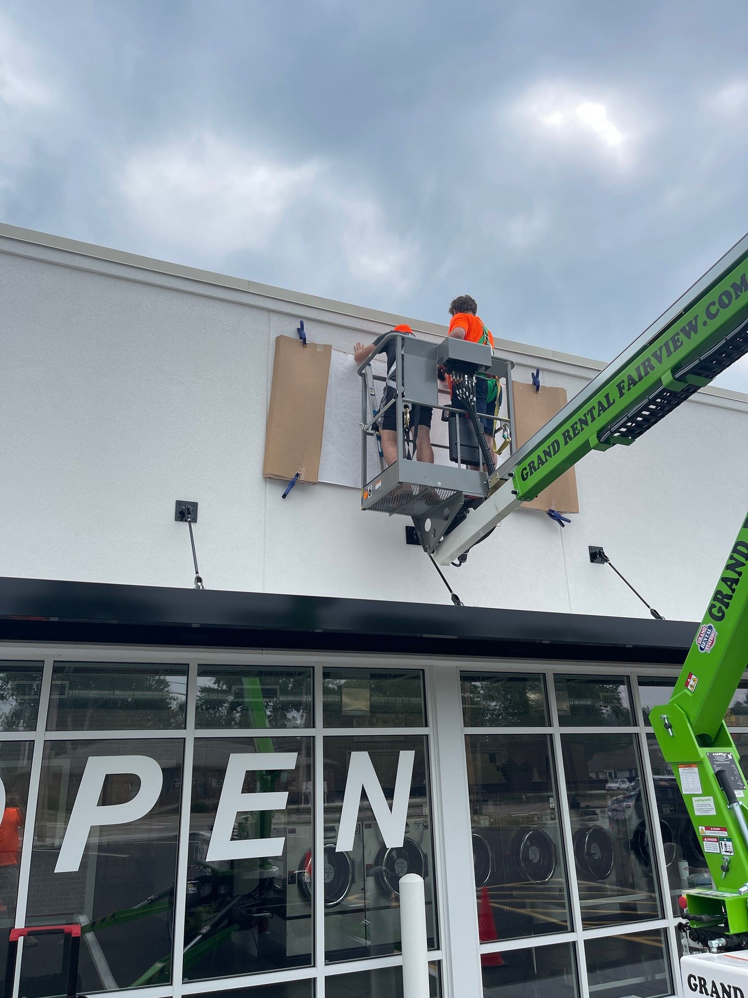 Two workers in an aerial lift installing a sign on a white building above a glass window displaying the word 