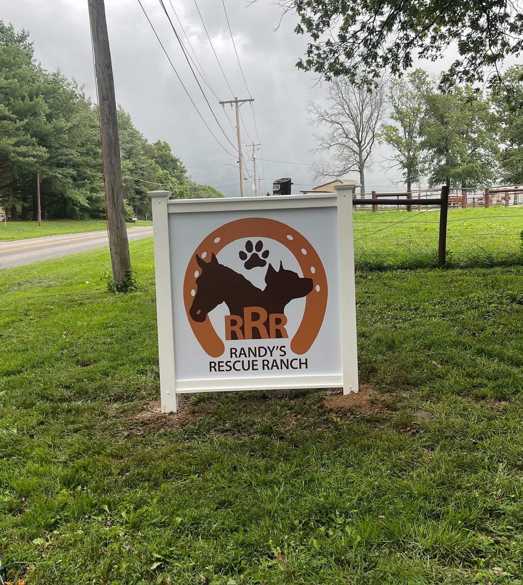 Sign for Randy's Rescue Ranch with horse, dog paw, horseshoe logo. White frame on a grassy lawn with trees in the background.