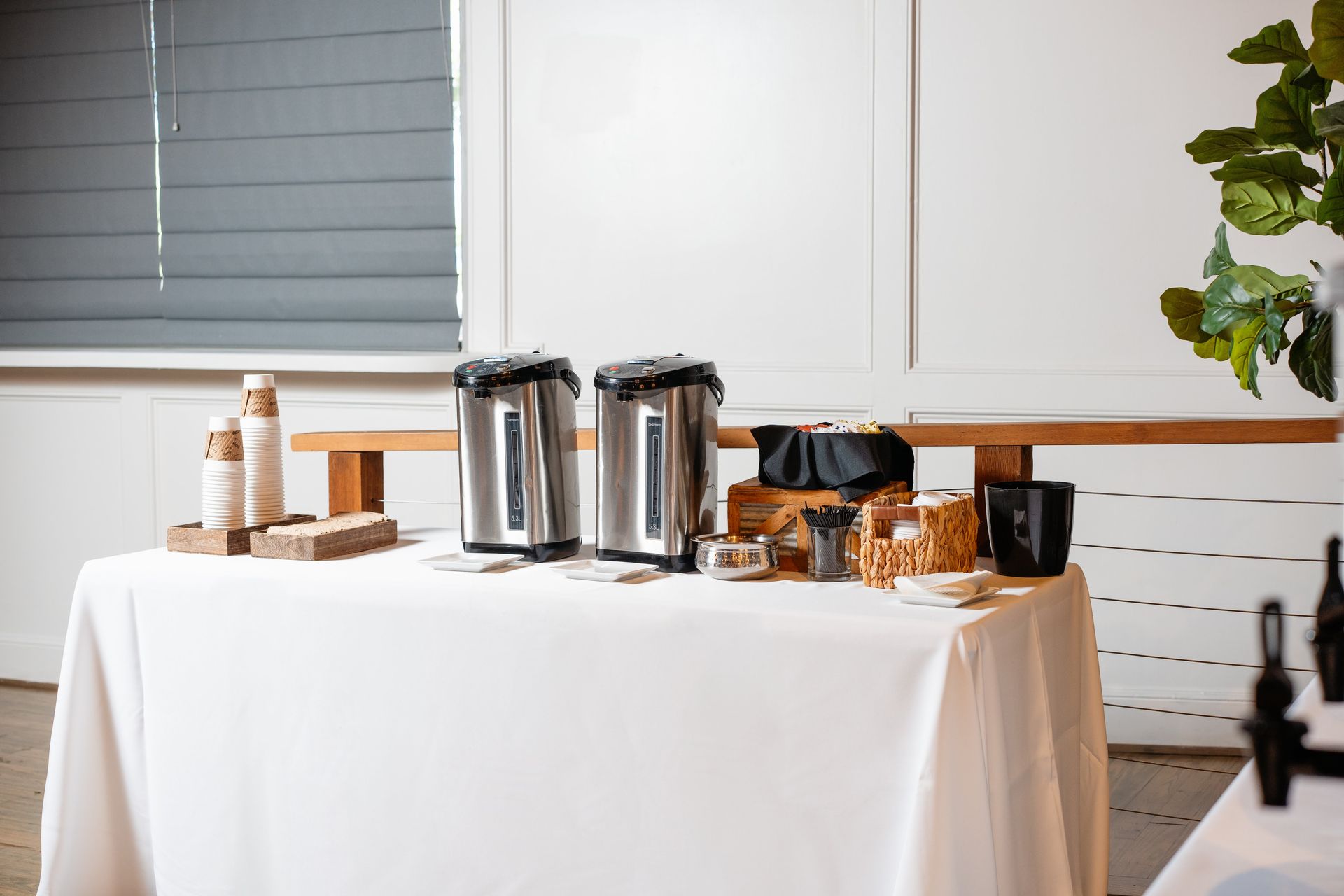 A table with a white tablecloth and coffee pots and cups on it.