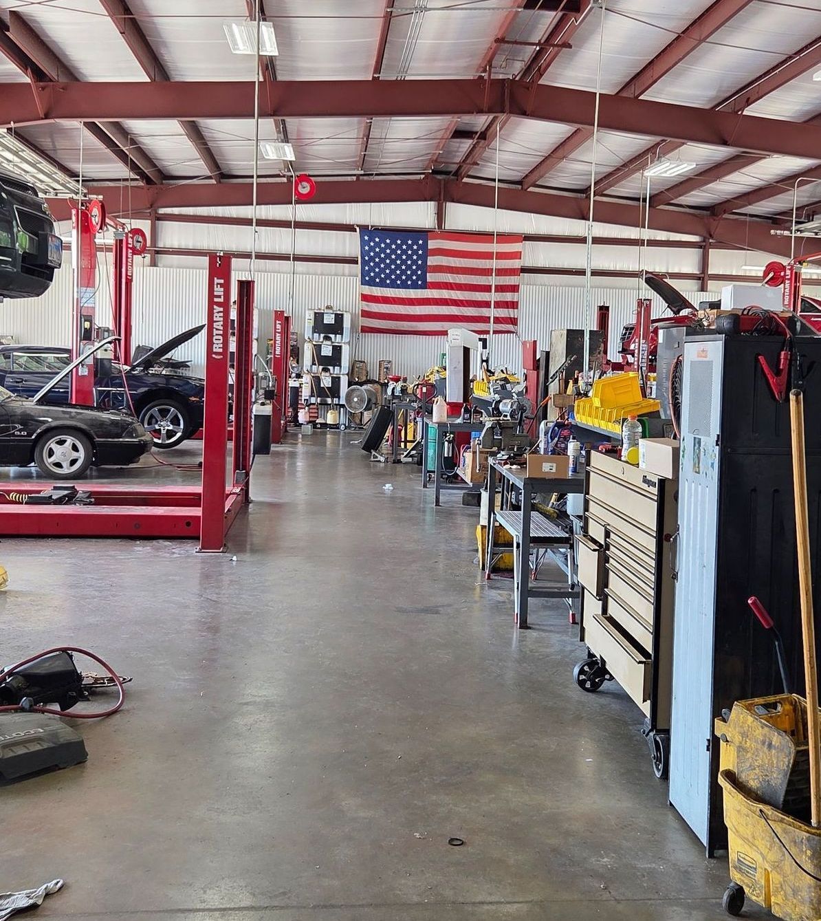 Interior of a car repair shop. Several cars are on lifts. Tools and workbenches are visible. An American flag hangs on the back wall.