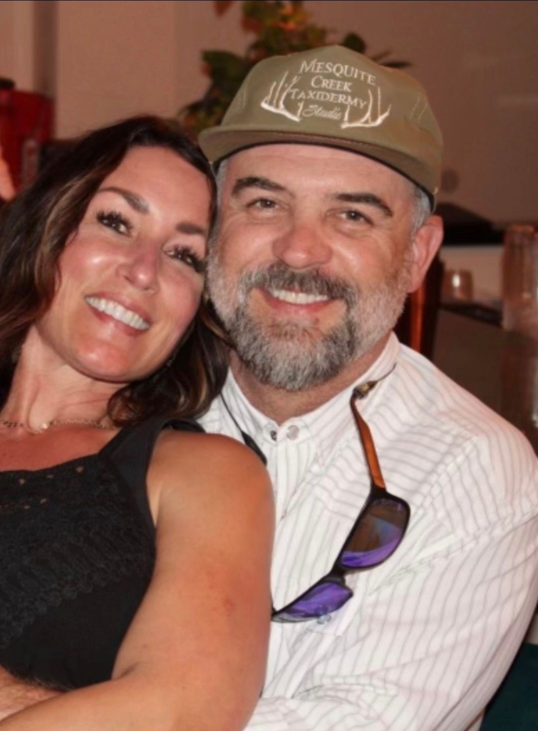 Woman and man smiling close together, outdoors; man in cap, holding sunglasses.
