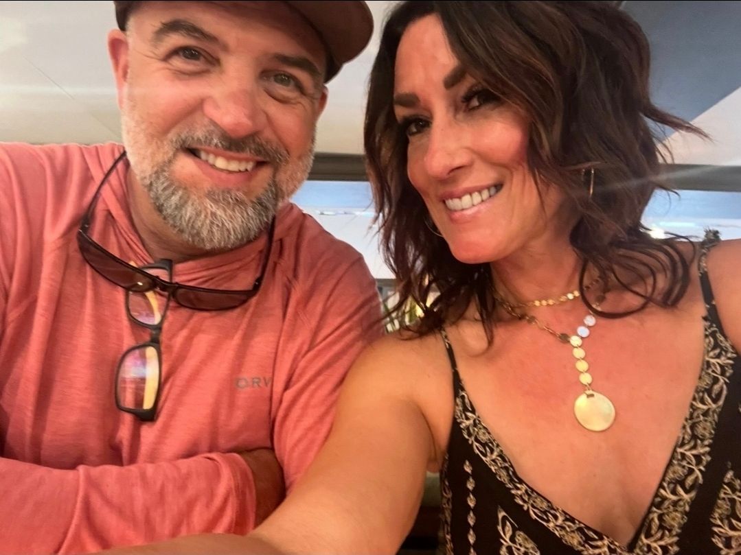 Man and woman smiling at the camera; man wears hat, sunglasses; woman wears necklace; close-up selfie.