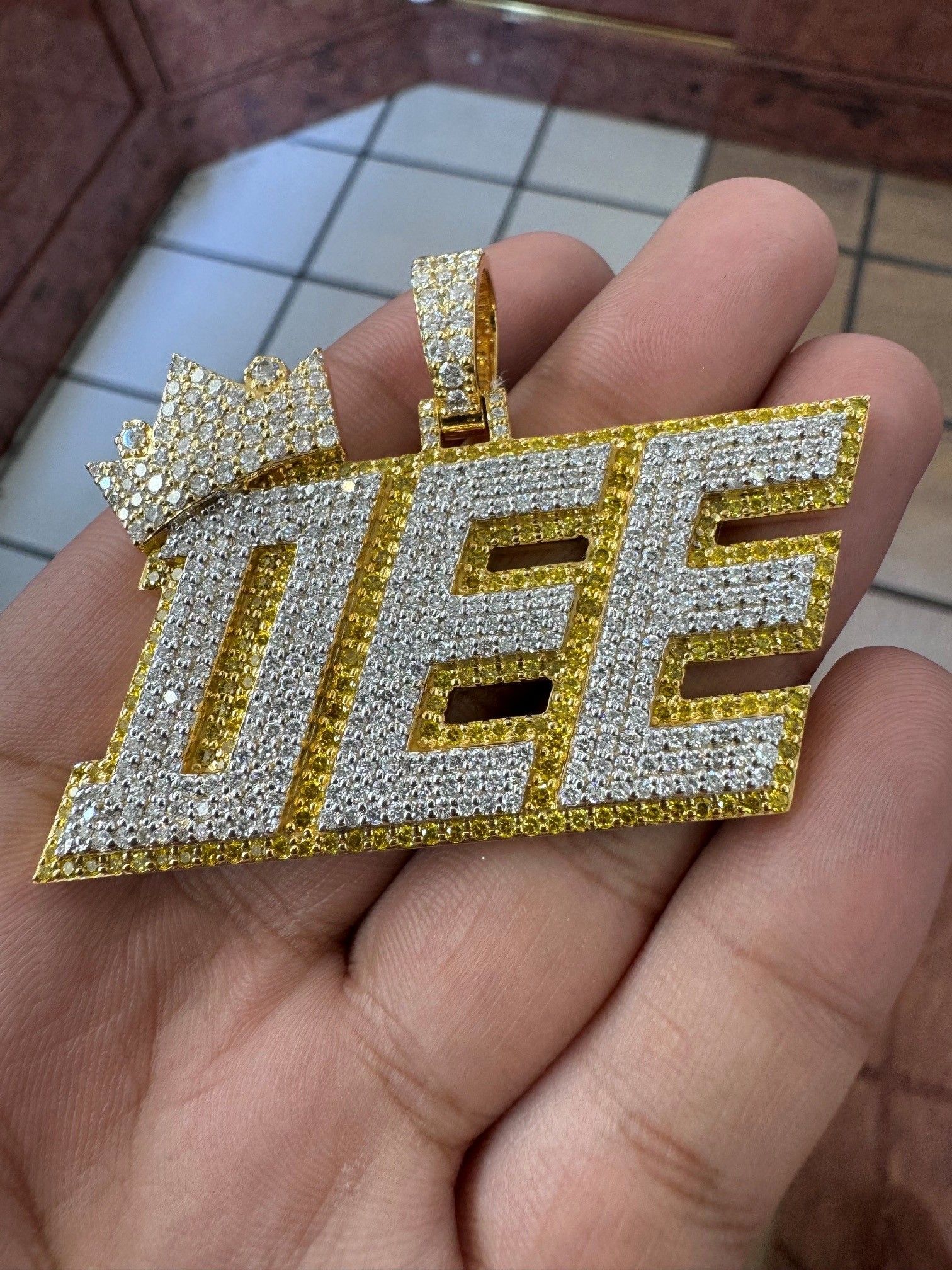 Gold, diamond-studded “DEE” pendant held in a hand over a tiled floor.