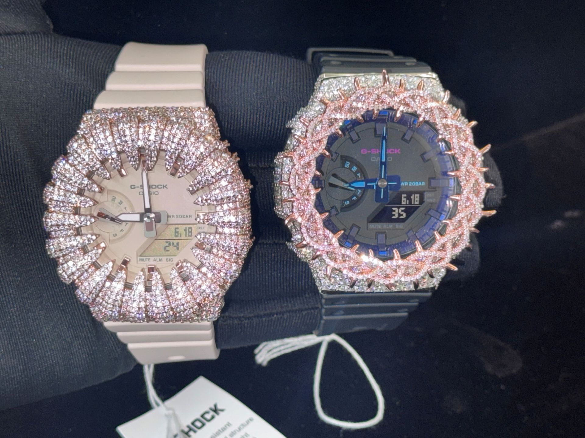 Two jeweled wristwatches on a dark seat, one cream and one black with pink accents.