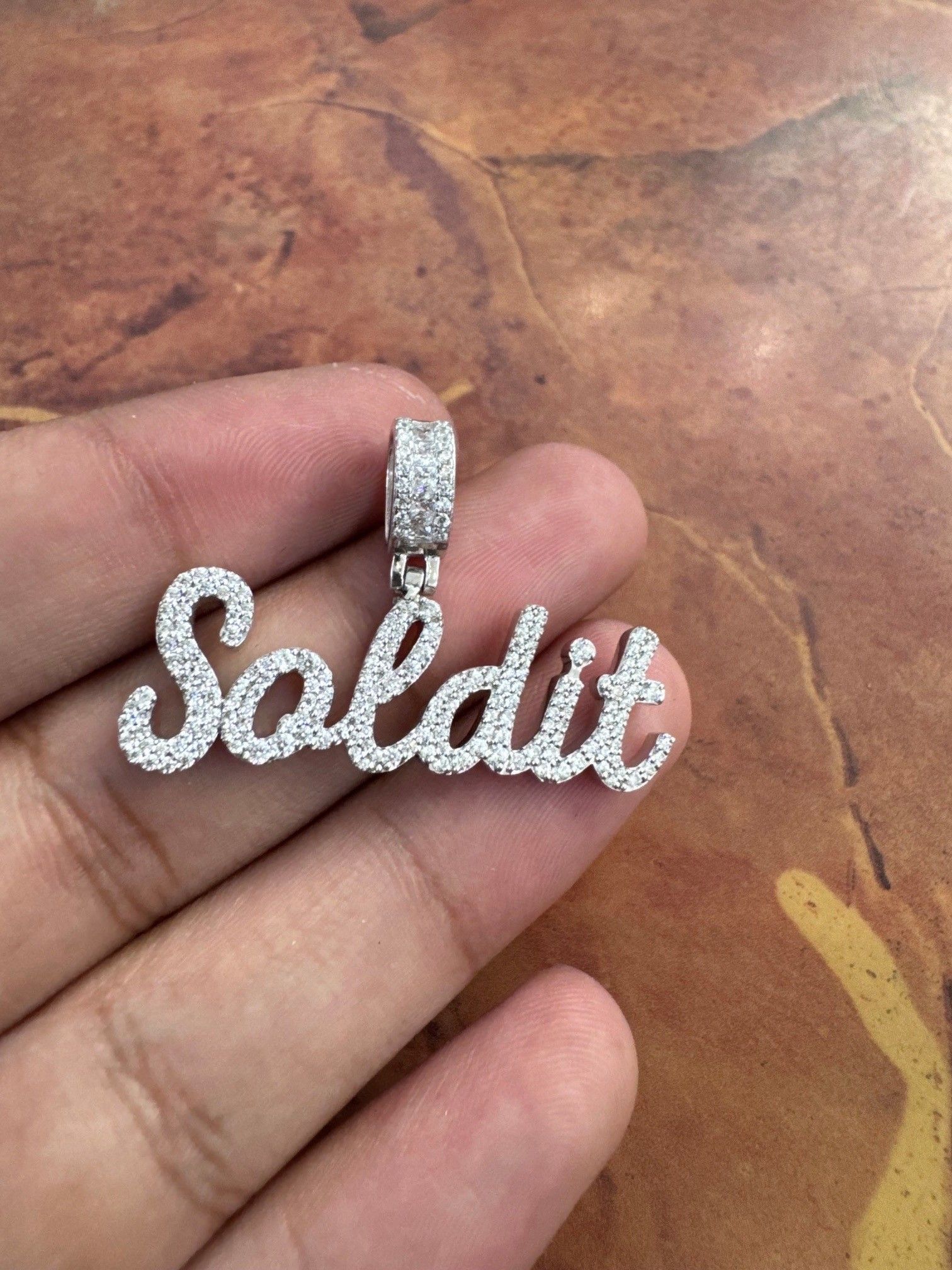 Silver script pendant spelling “Sabbitt” held in a hand over a brown stone surface