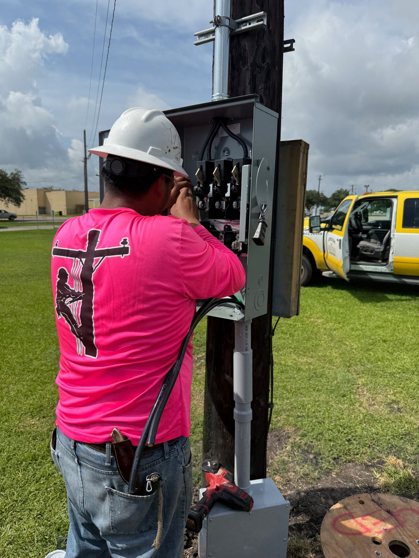 A utility worker in a pink shirt and hard hat installs wiring in an outdoor electrical box on a utility pole.