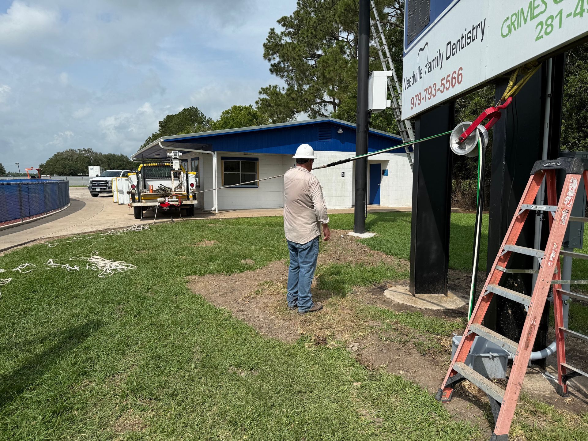 A person in a hard hat inspects a sign installation site with a ladder and a utility vehicle in the background.