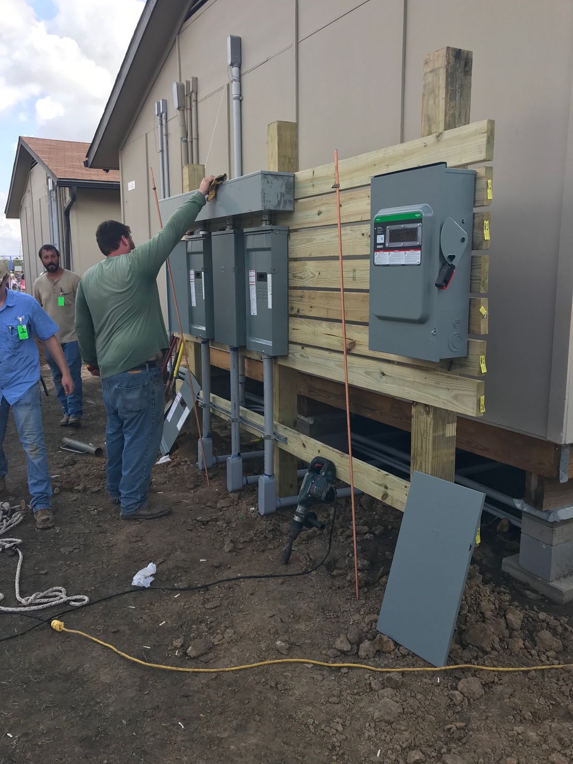Workers installing electrical panels mounted on a wooden frame against the exterior wall of a building.