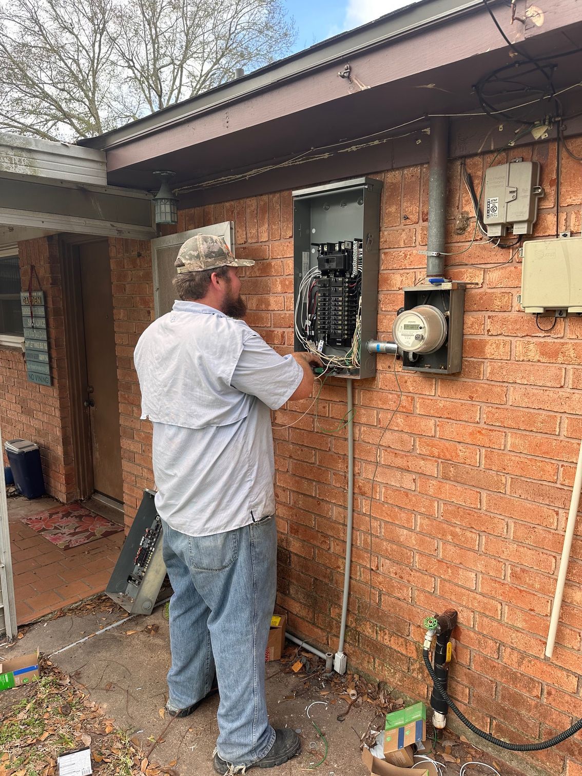 A person in a light-colored shirt and cap working on an open electrical service panel on an exterior brick wall.