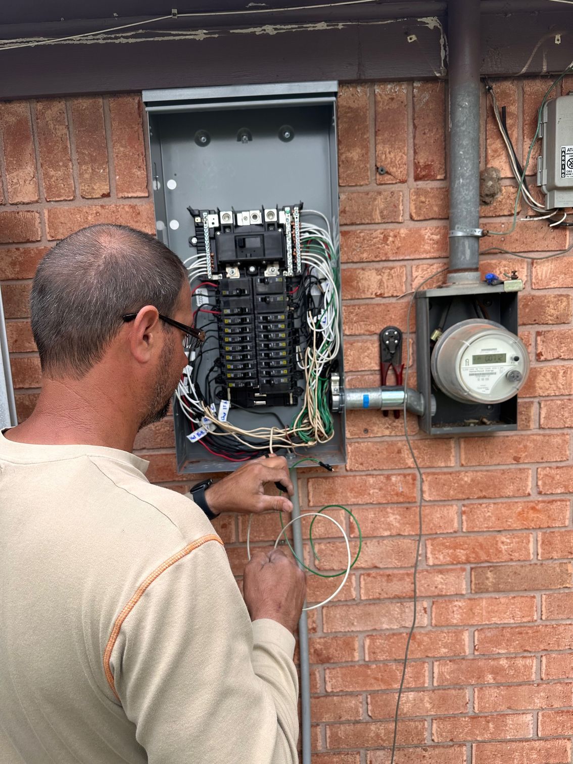 A person works on an open electrical breaker panel mounted on a brick exterior wall next to an electric meter.