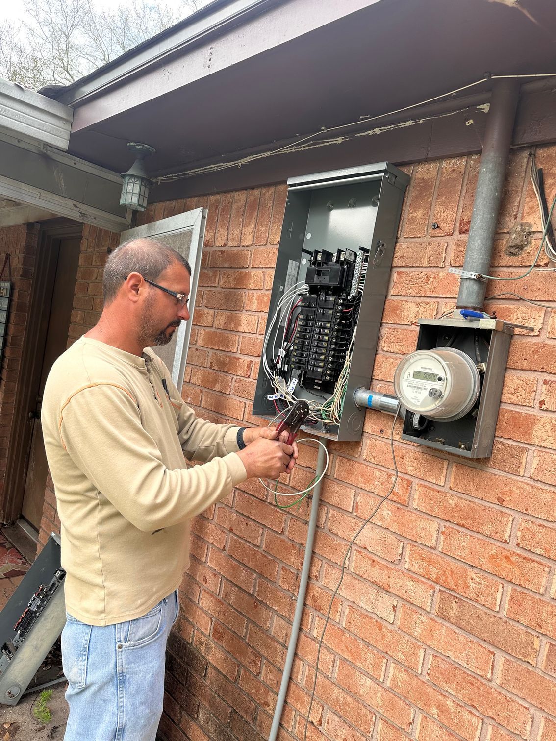 Electrician working on an open electrical service panel mounted to a brick wall next to an electric meter.
