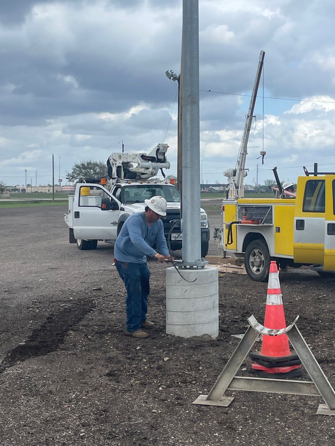 A worker in a hard hat and blue long-sleeved shirt installs a metal pole on a concrete base at an outdoor worksite.