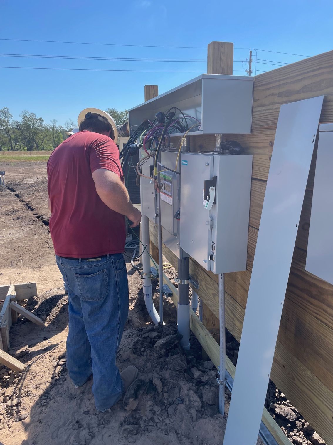 A person in a maroon shirt and hard hat installs wiring into outdoor electrical boxes mounted on a wooden fence.