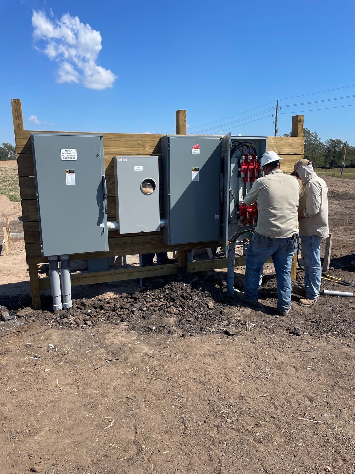 Two technicians in hard hats work on an open electrical service panel mounted on a wooden frame in a rural field.