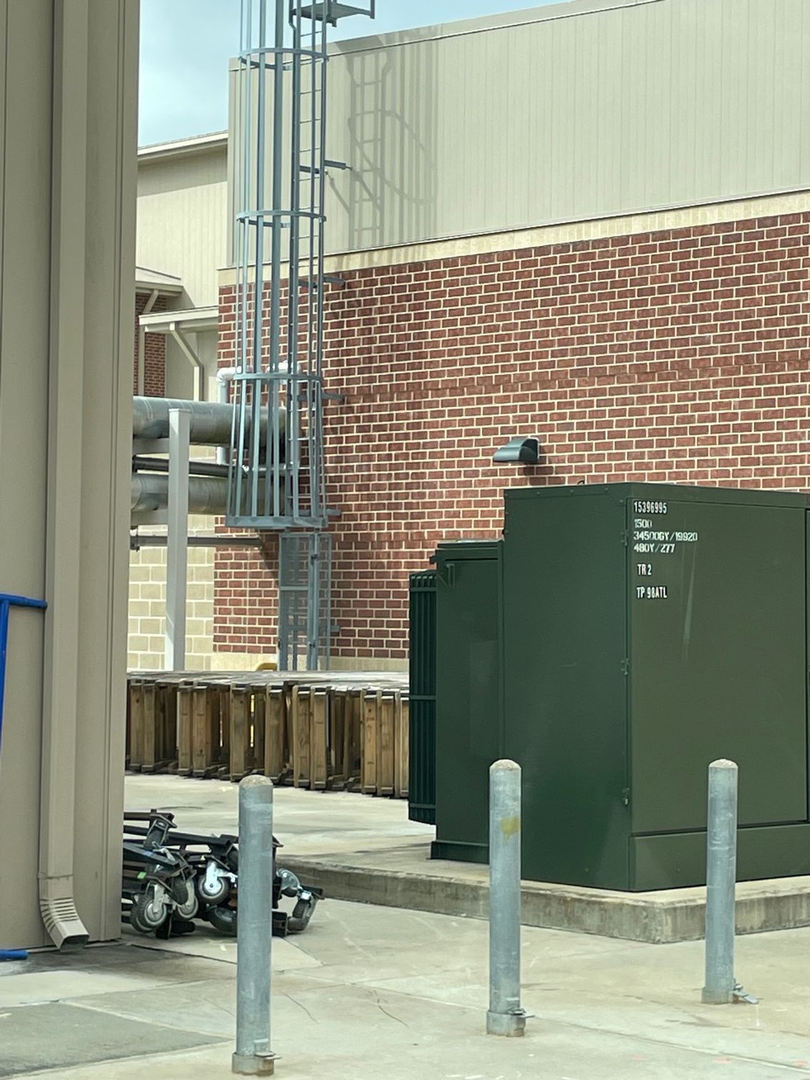 Brick building with a metal ladder, pipes, and electrical boxes. Metal bollards in front.