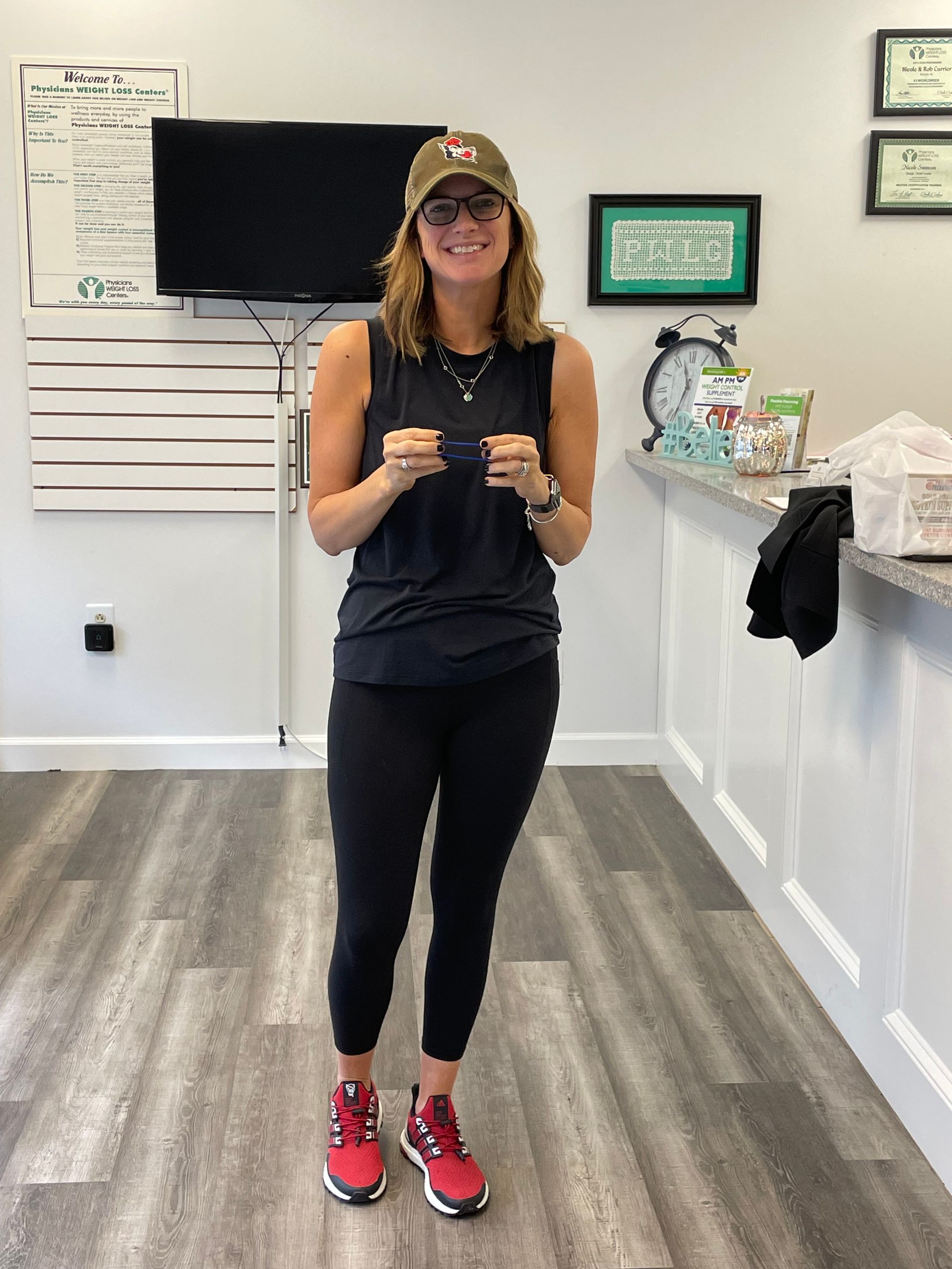 Woman in cap and glasses, holding glasses, standing indoors, wearing black workout clothes and red shoes.