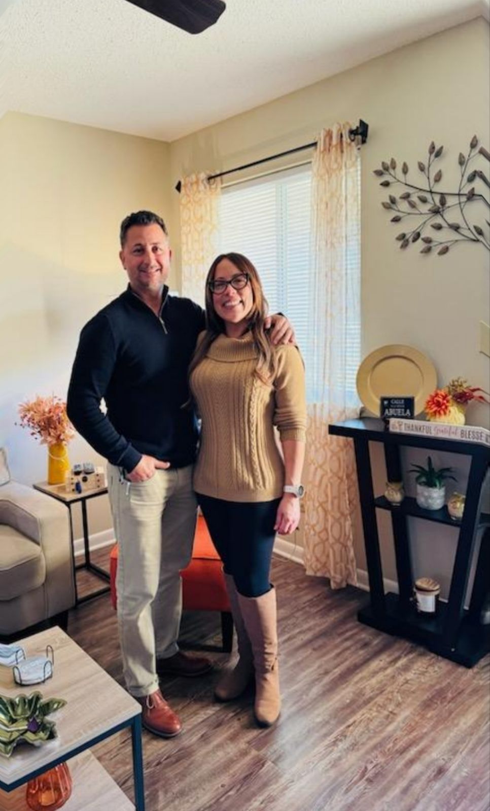 Couple smiling, posing together inside a well-lit living room. Man in black sweater and khakis, woman in tan sweater and skirt.