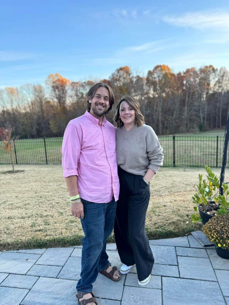 Couple poses outdoors; man in pink shirt, jeans, sandals, woman in sweater, black pants, white sneakers; backyard, autumn trees.
