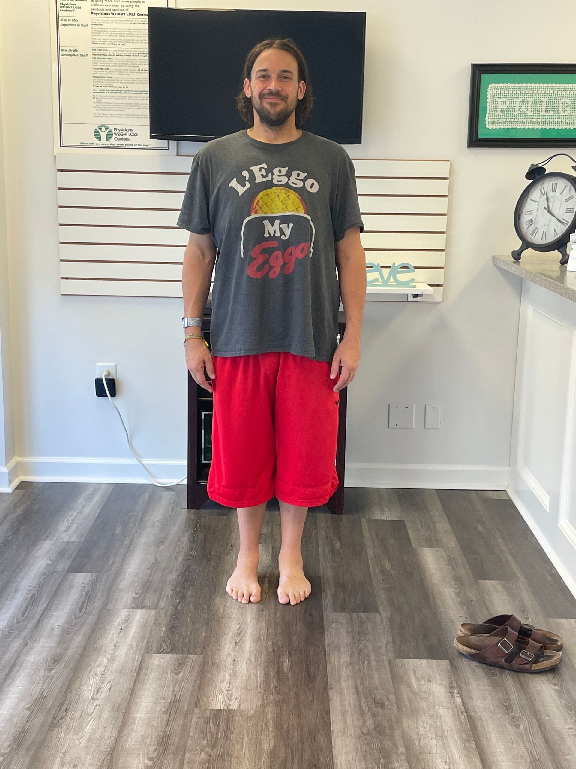 Man standing indoors, wearing a gray t-shirt and red shorts, barefoot on a wood floor.