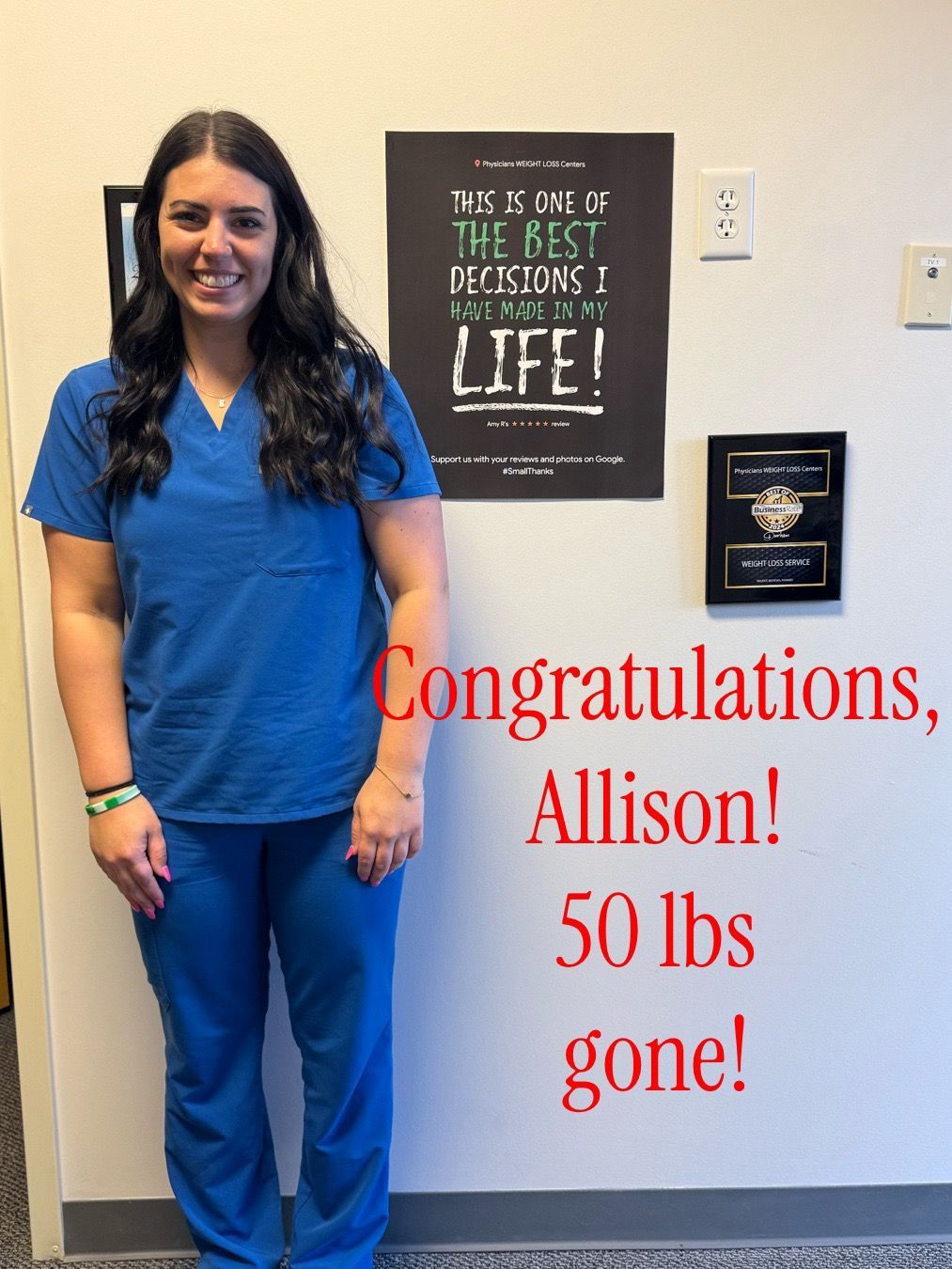 Woman in blue scrubs smiles, standing in front of a wall with a sign. Text says 